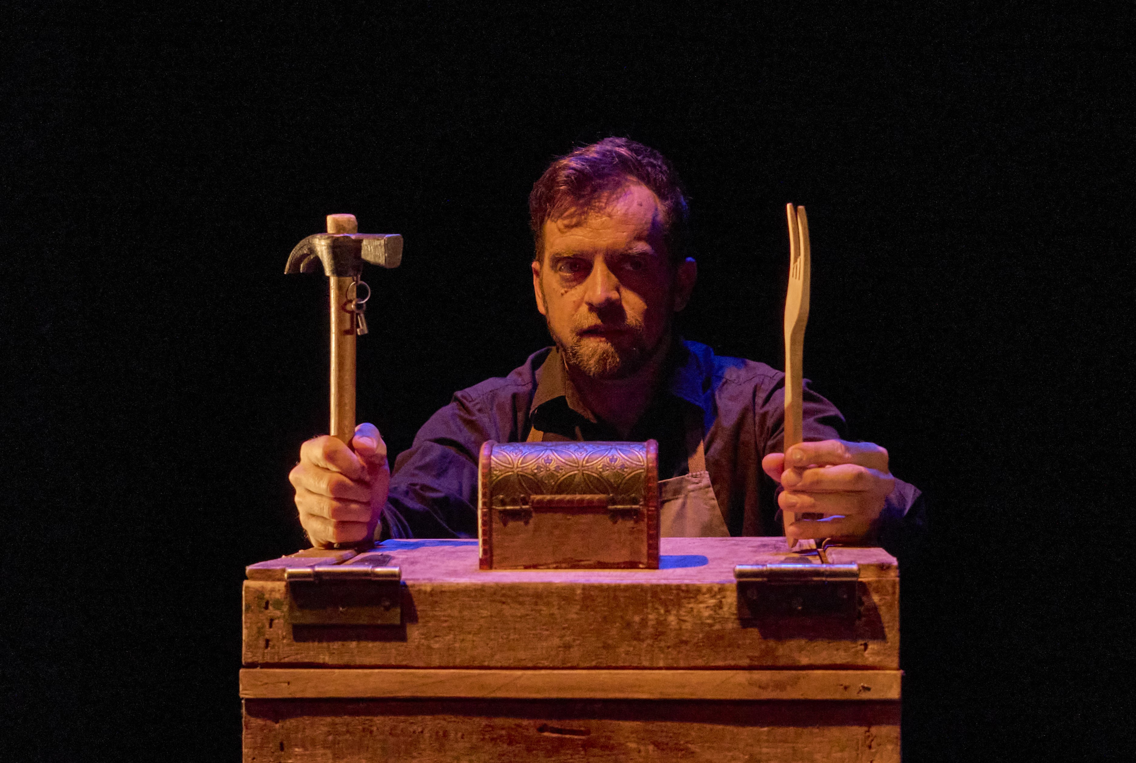 Un actor con delantal, detrás de una caja de madera, sostiene un martillo con unas llaves colgando en una mano y un tenedor de madera en la otra. Delante de él, sobre la caja, hay un pequeño cofre de metal labrado. Fondo negro y luz cálida.