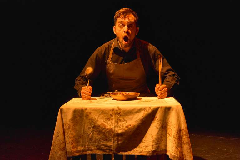 El actor, sentado a una mesa con mantel de flores y la boca muy abierta, sostiene una cuchara y un tenedor de madera en alto, a punto de lanzarse sobre un cuenco de comida.
