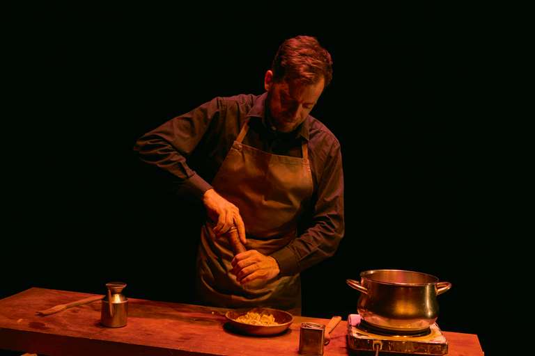 El actor, con delantal, muele pimienta sobre un cuenco de madera en una mesa de trabajo. Junto a él hay una aceitera, un tenedor de madera y una olla sobre un hornillo eléctrico.