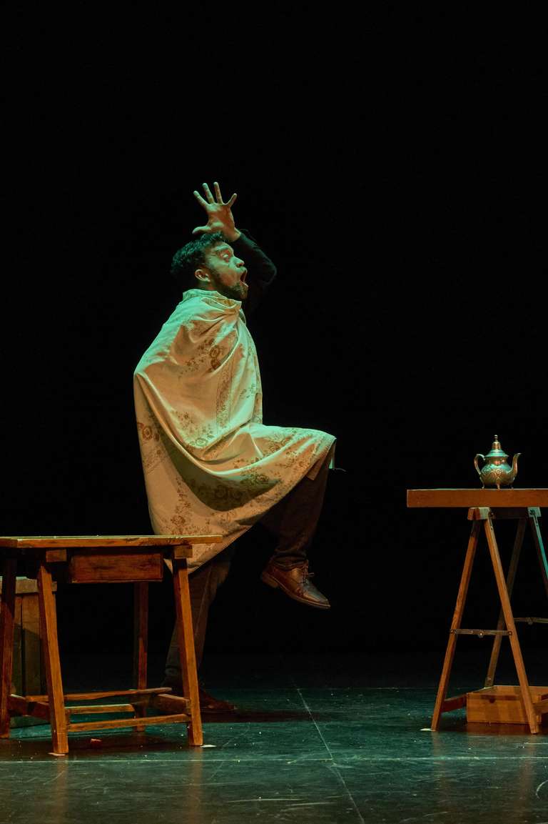 El actor, cubierto con un mantel de flores a modo de capa, levanta una rodilla y una mano en alto con gesto teatral de sorpresa. Está entre dos mesas de madera, y sobre la de la derecha hay una tetera plateada.