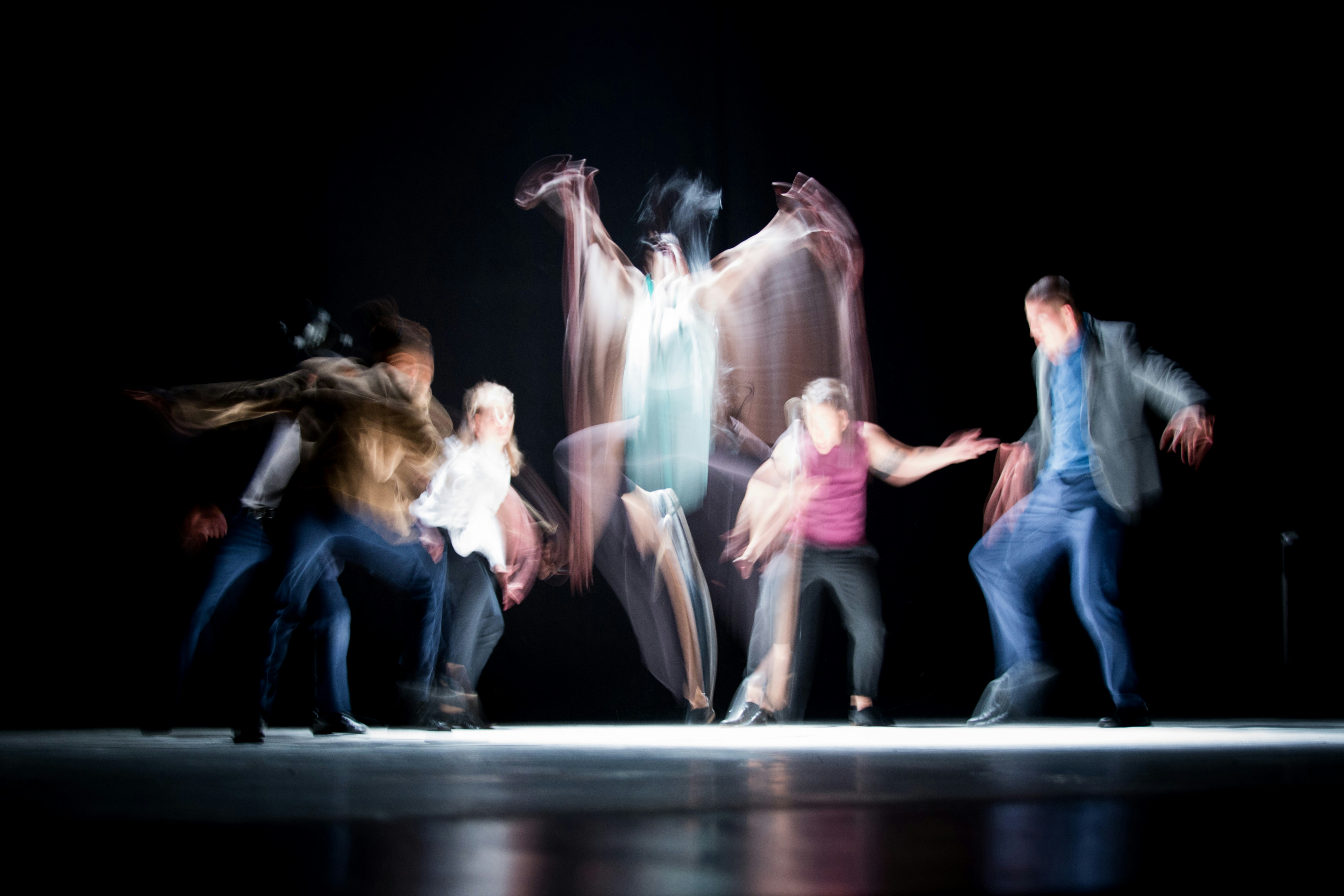 Grupo de bailarines en un escenario oscuro, fotografiados con larga exposición: sus movimientos dejan estelas de color y, en el centro, una figura salta con los brazos abiertos como si fueran alas.