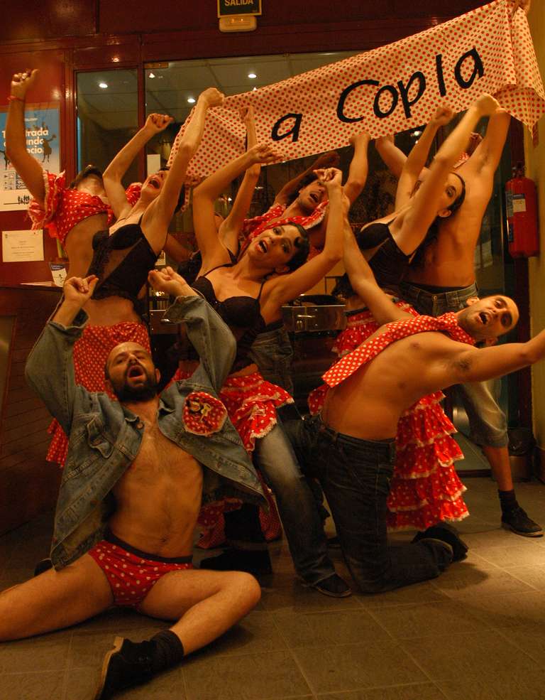 La compañía posa en el vestíbulo de un teatro con volantes y pañuelos de lunares rojos y blancos, brazos en alto y cara de cantar a pleno pulmón, bajo una pancarta de lunares que dice «a Copla».