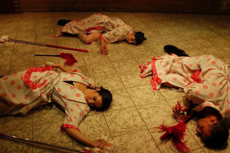 Tres intérpretes con batas de lunares rosas y delantales con volantes yacen agotados en un suelo de baldosas, rodeados de plumeros rojos, una fregona y pulverizadores de limpieza.