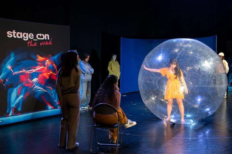 Una bailarina con vestido amarillo baila dentro de una gran esfera transparente en el escenario, mientras varias personas del público la observan, de pie o sentadas. A la izquierda, una pantalla proyecta la imagen de «stage_on the road».