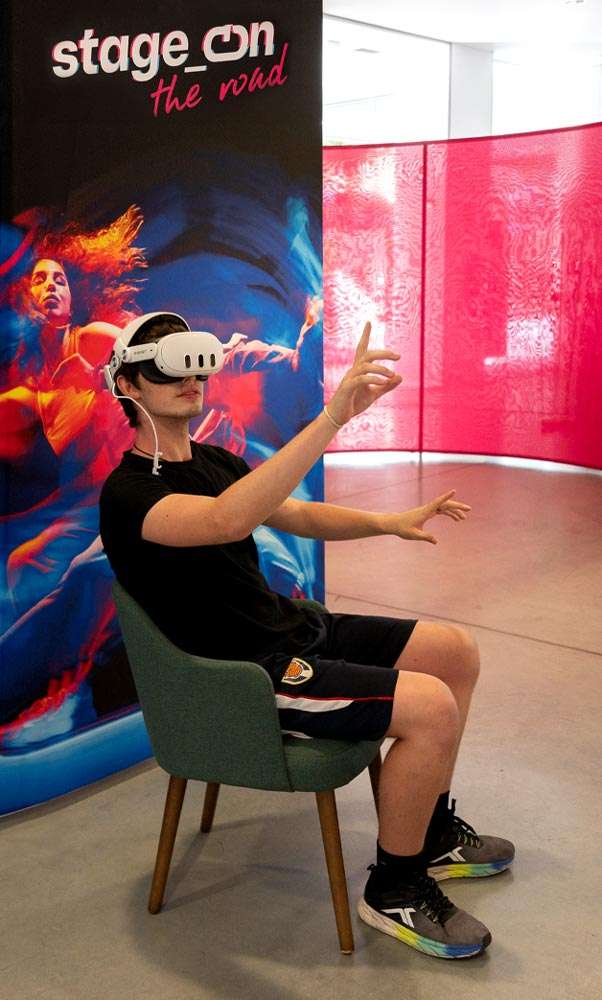 Un joven sentado en un sillón verde, con gafas de realidad virtual, extiende los brazos hacia delante como si tocara algo invisible. Detrás hay un cartel de «stage_on the road» con una bailarina en tonos rojos y azules, y paneles de tela rosa.