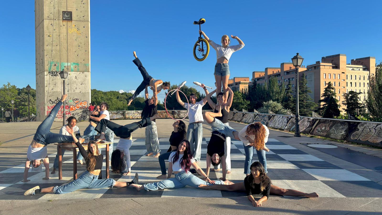 Una quincena de estudiantes de circo posan al aire libre sobre un suelo de cuadros blancos y negros, en un día soleado. Hacen spagats, verticales y portés: una chica de pie sobre los hombros de otra sostiene un monociclo en alto, otra vuela en un portés acrobático y un chico levanta mazas de malabares junto a un aro. Al fondo, una torre de escalada con grafitis y edificios de ladrillo.