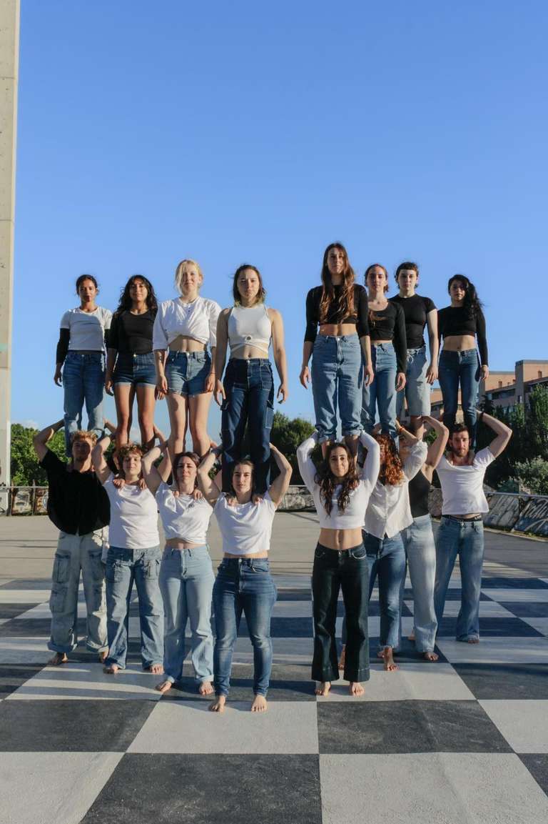 Dieciséis estudiantes de circo forman una torre humana de dos alturas sobre un suelo de cuadros blancos y negros. Ocho están de pie sobre los hombros de otros ocho, que los sujetan por los tobillos. Todos van descalzos, con vaqueros y camisetas blancas o negras, bajo un cielo azul.