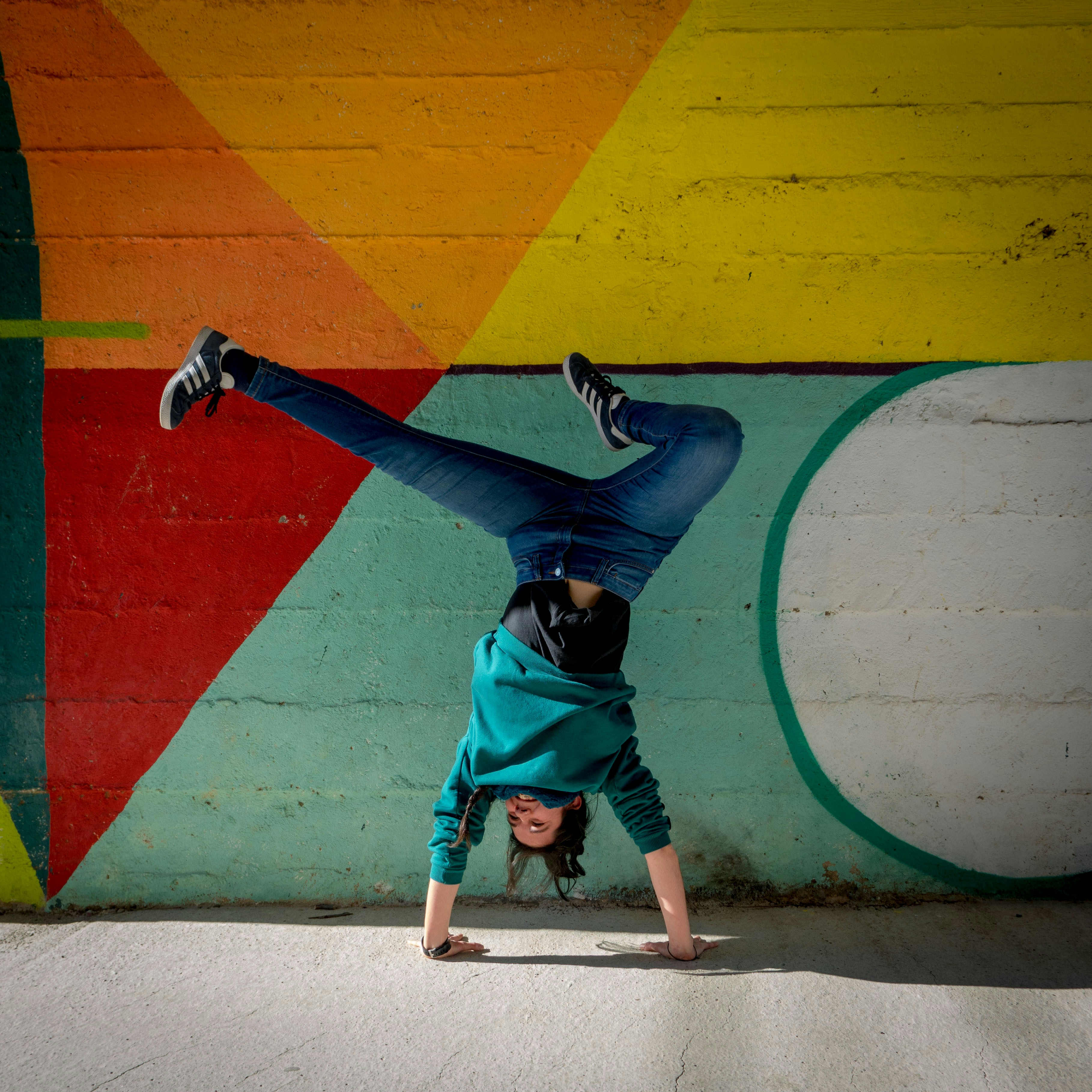 Una joven con sudadera turquesa y vaqueros hace el pino, con las piernas abiertas, delante de una pared pintada con grandes formas geométricas en rojo, naranja, amarillo y verde agua.