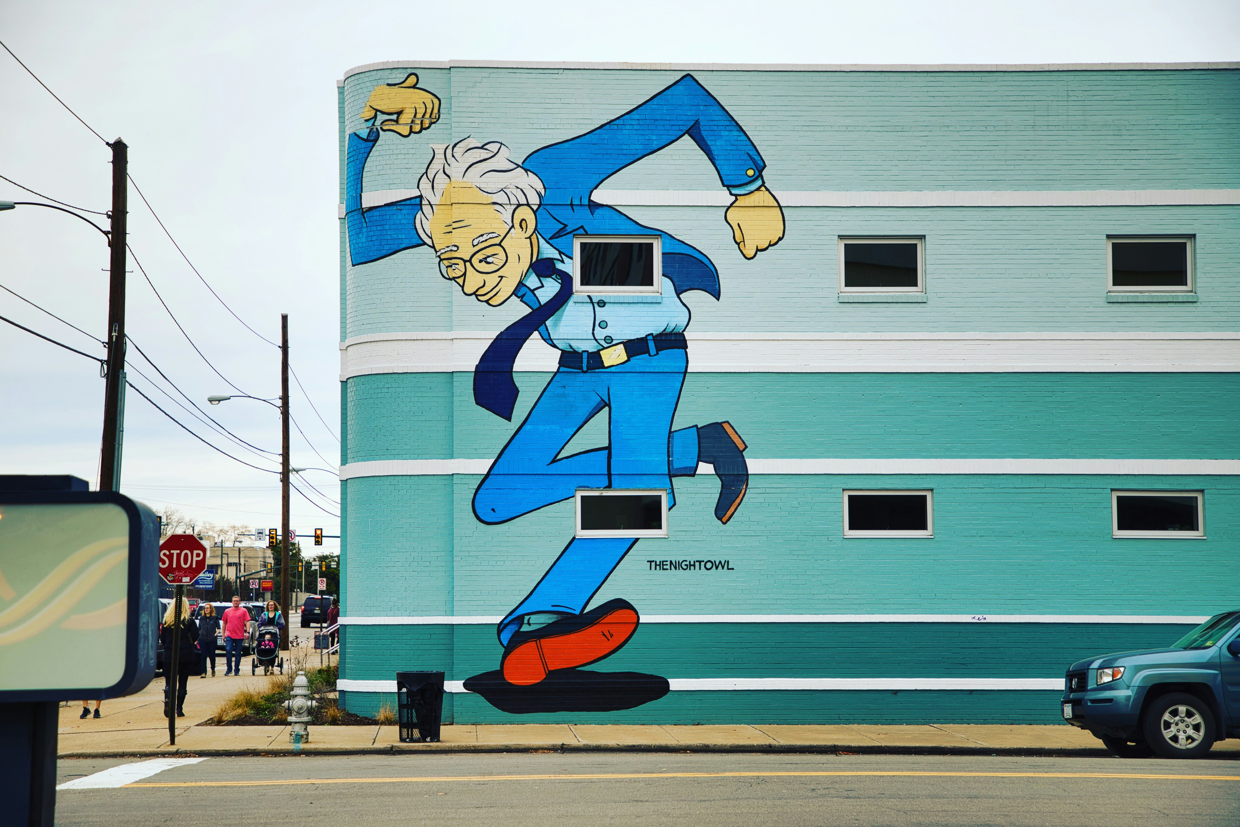 Mural de estilo cómic en la fachada turquesa de un edificio: un hombre mayor de pelo blanco, gafas y traje azul avanza a grandes zancadas, con energía, junto a una calle con semáforos y peatones.