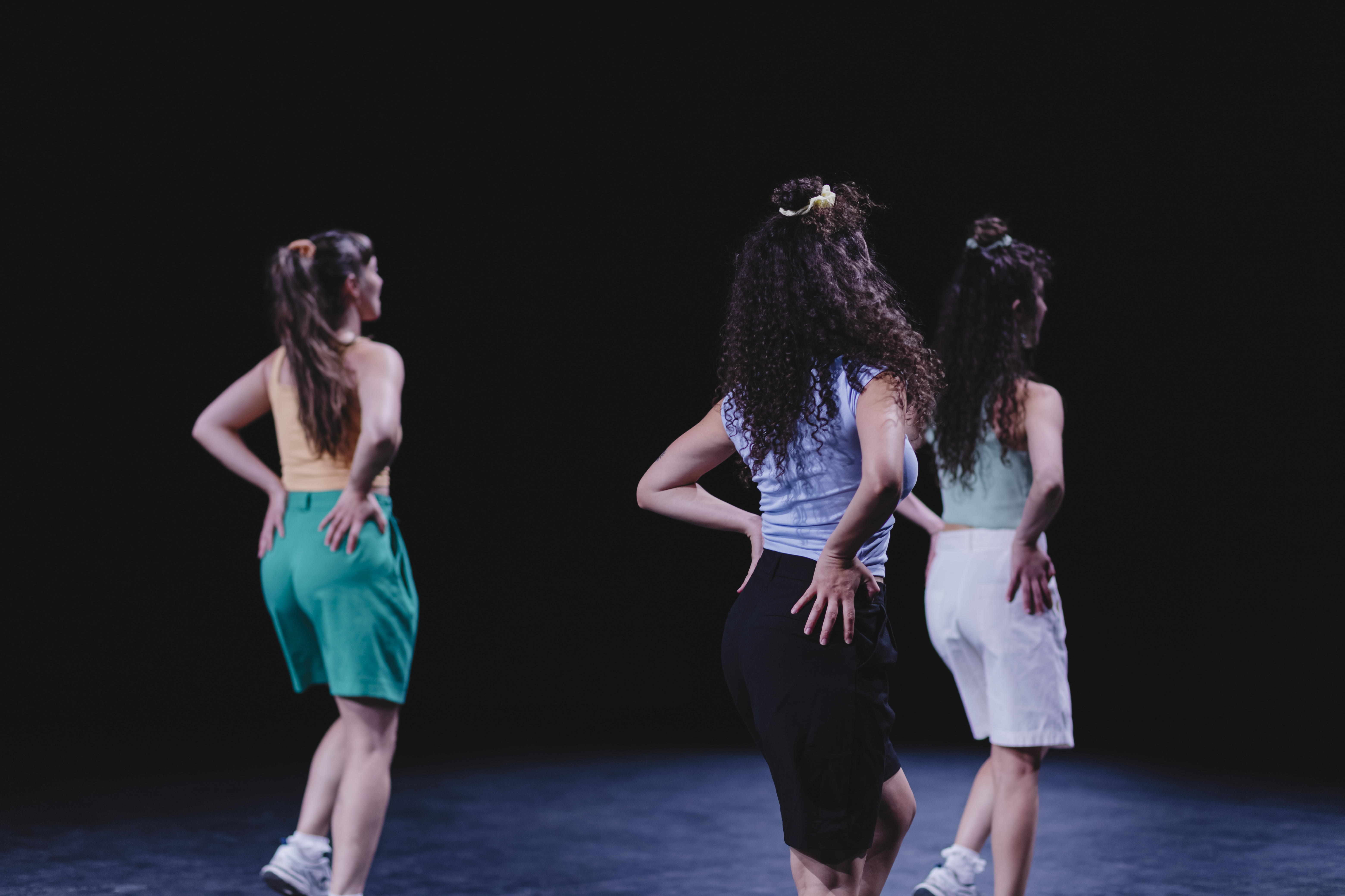 Tres bailarinas de espaldas, con las manos en las caderas, se mueven al unísono sobre un escenario oscuro. Llevan camisetas de tirantes y bermudas de colores, y el pelo recogido con coleteros.
