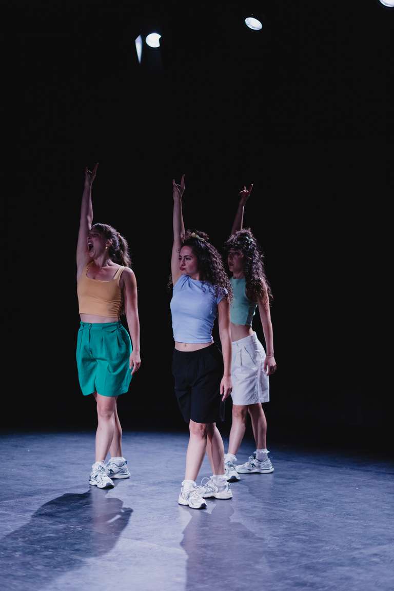 Tres bailarinas en fila sobre un escenario oscuro levantan el brazo derecho con la mano en forma de cuernos. La primera canta con la boca muy abierta. Llevan tops de colores, bermudas y zapatillas blancas.