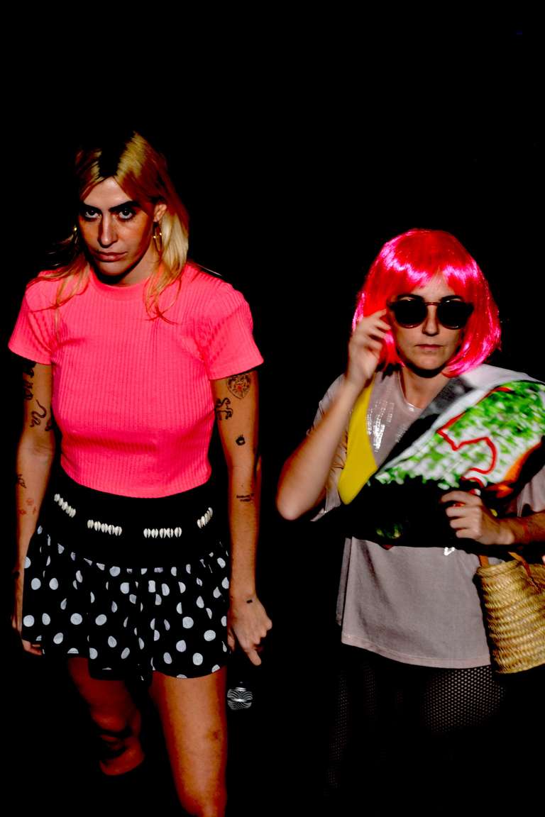 Dos intérpretes miran a cámara con gesto serio sobre fondo negro. A la izquierda, una con camiseta rosa flúor y falda de lunares sostiene un micrófono. A la derecha, otra con peluca rosa y gafas de sol lleva una toalla de playa al hombro y un capazo.